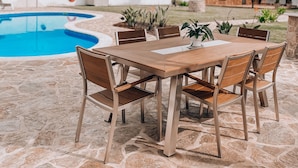 An outdoor patio with a stone floor, a wooden table with chairs, and a swimming pool in the background.