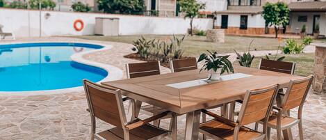 Un patio exterior con piso de piedra, una mesa de madera con sillas y una piscina al fondo.