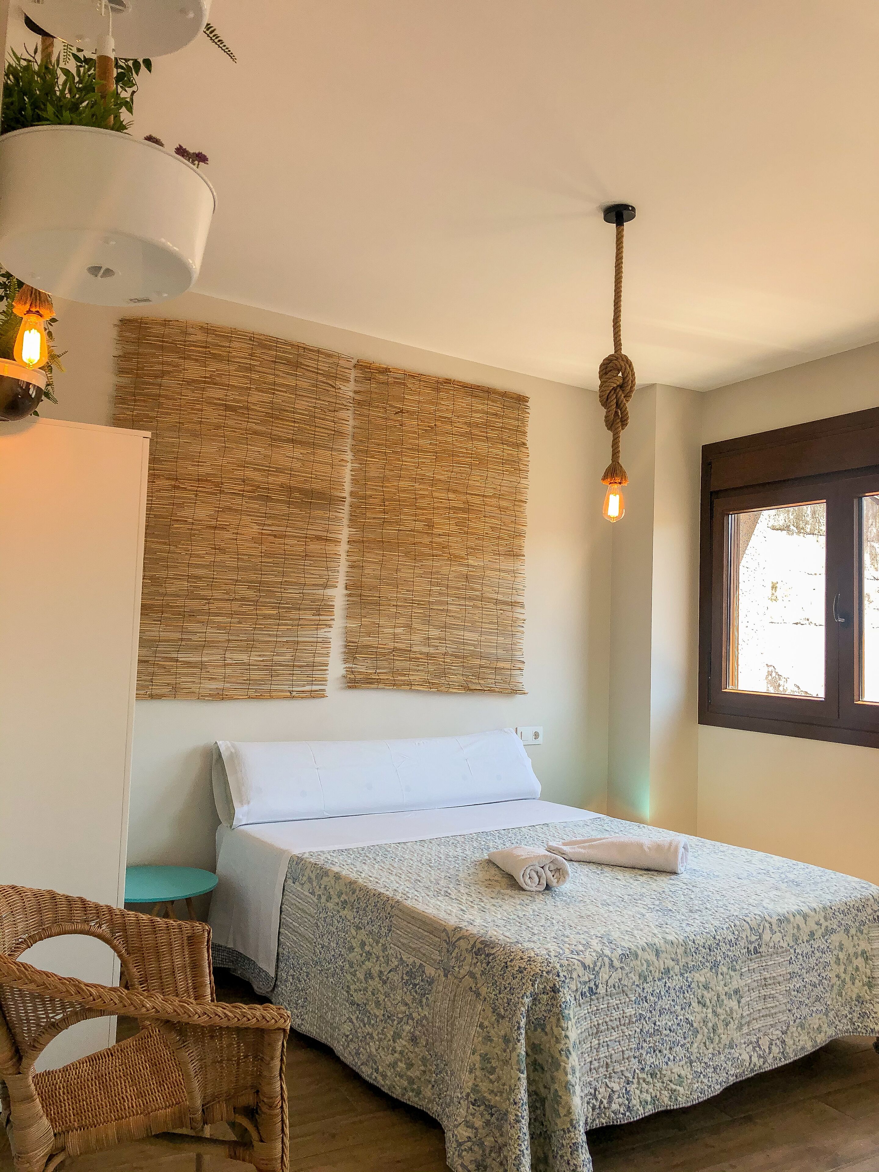 Un dormitorio con una cama, una silla de mimbre, una lámpara colgante y una ventana con vistas.