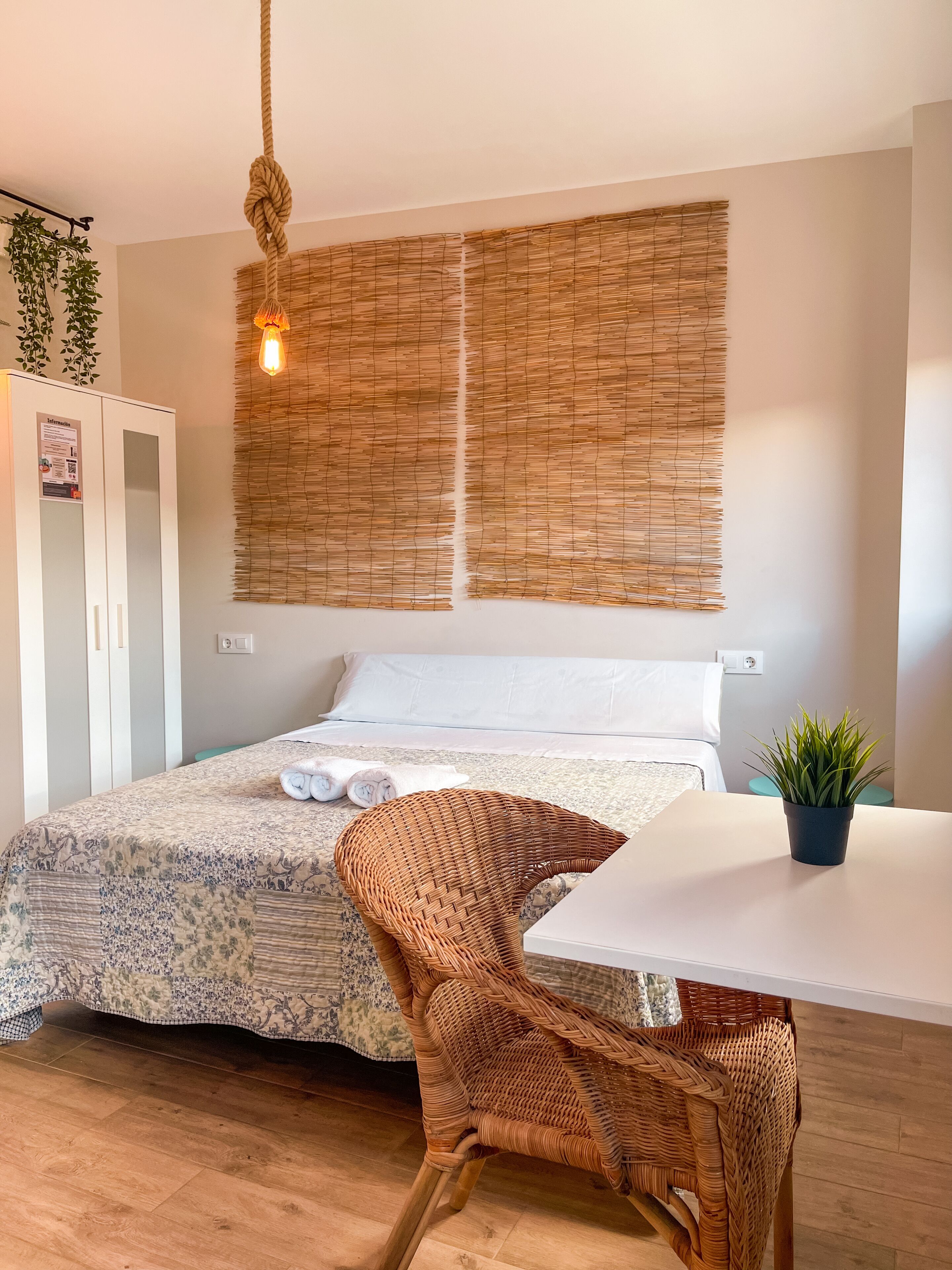 Un dormitorio ordenado con una cama, una silla de mimbre, una mesita y una lámpara colgante.