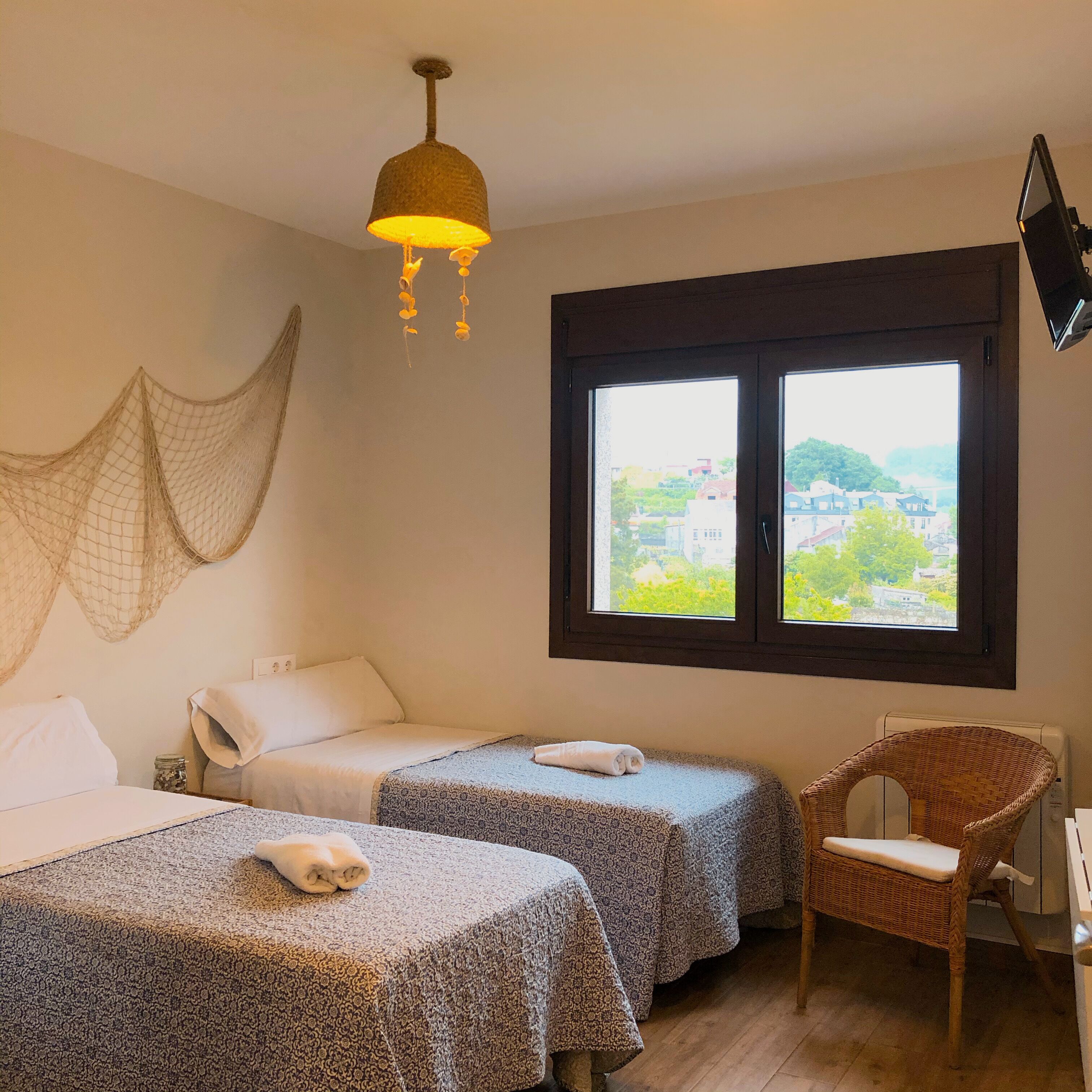Ein Zimmer mit zwei Betten, einem Korbstuhl, einem Fenster mit Ausblick und einer Pendelleuchte.