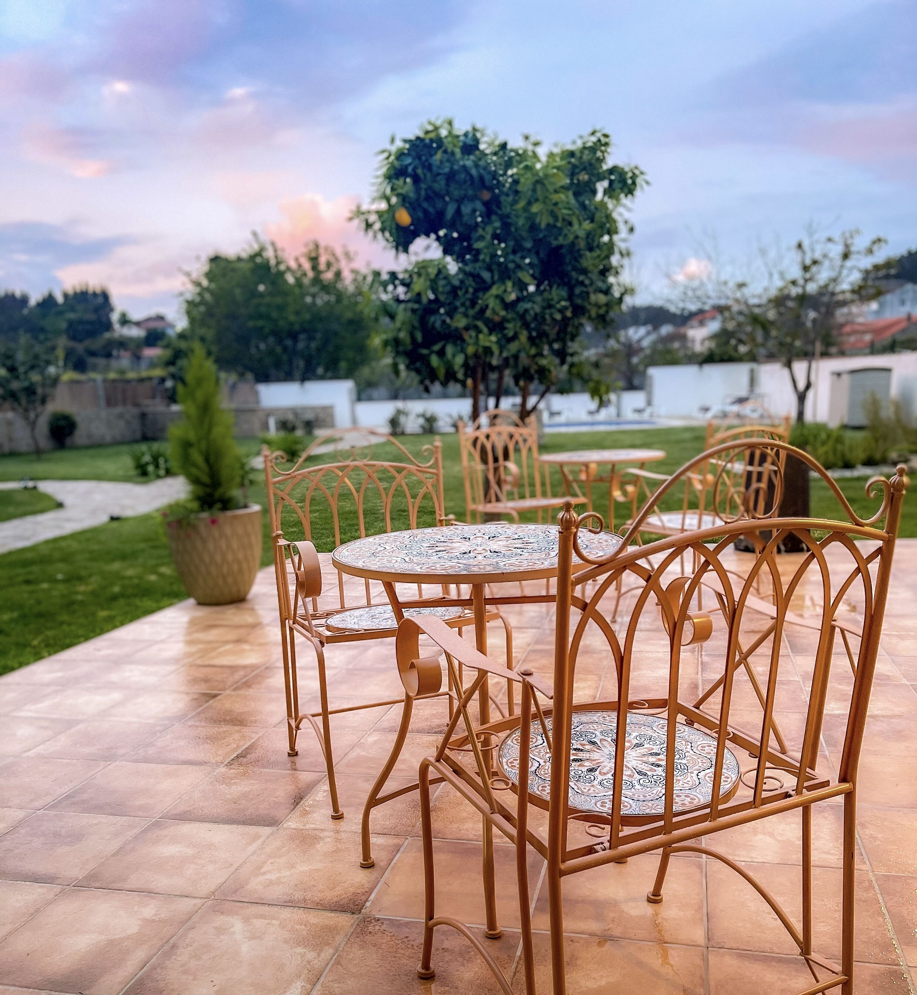 An outdoor patio with metal chairs and a round table, surrounded by a garden and a view of the sky.