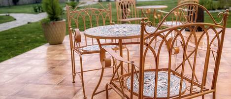 An outdoor patio with metal chairs and a round table, surrounded by a garden and a view of the sky.