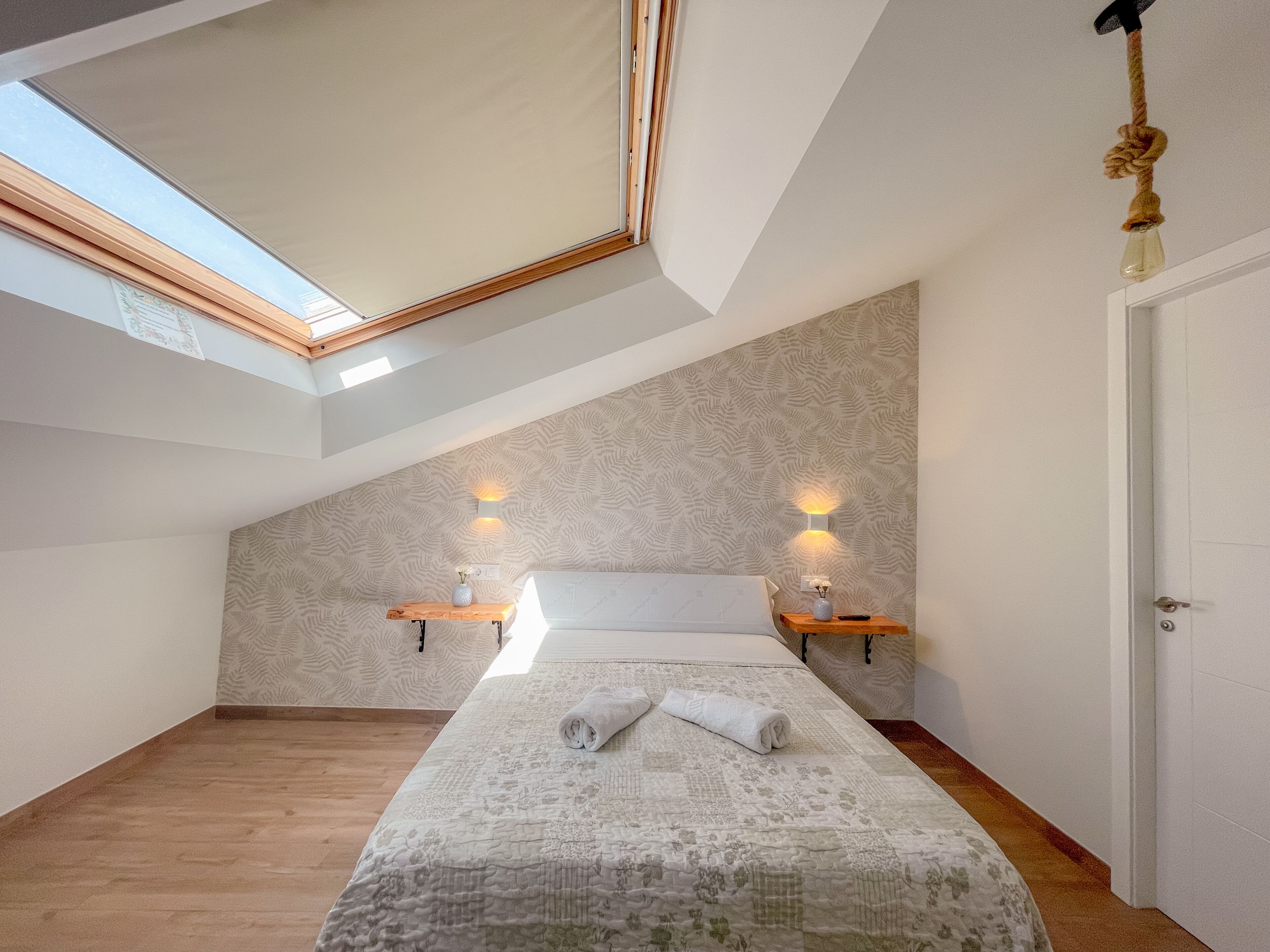 A bedroom with a sloped ceiling, skylight, two bedside tables with lamps, a bed with a rolled towel, and a door.