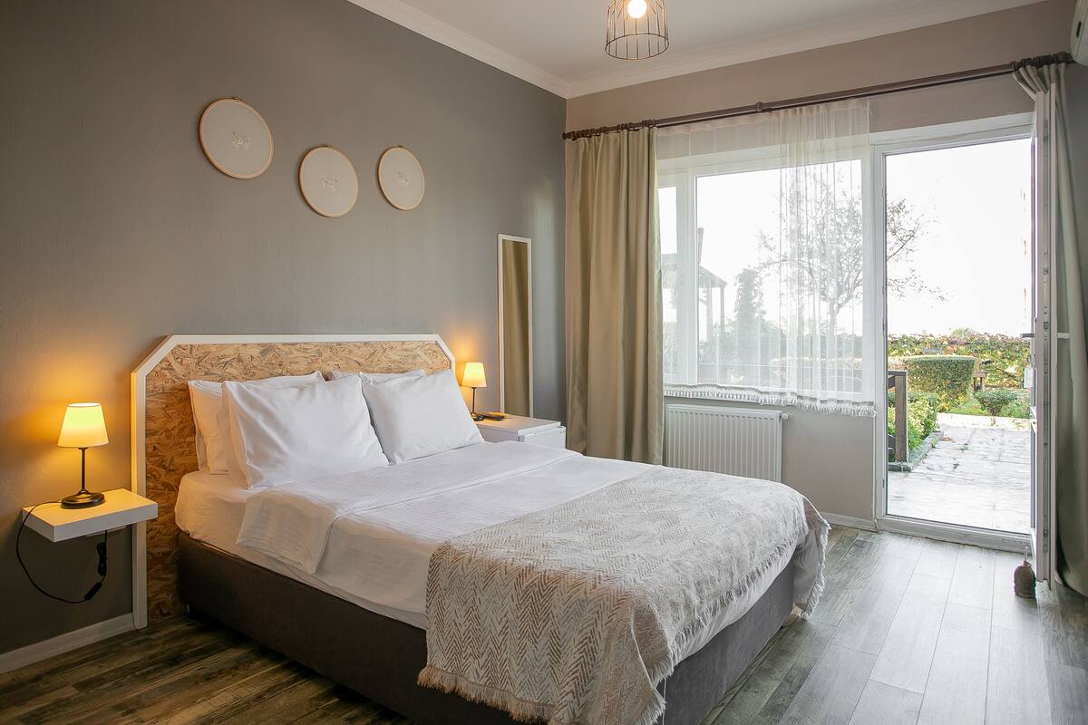 A bedroom with a bed, bedside lamps, a window with curtains, and a view of the outdoors.