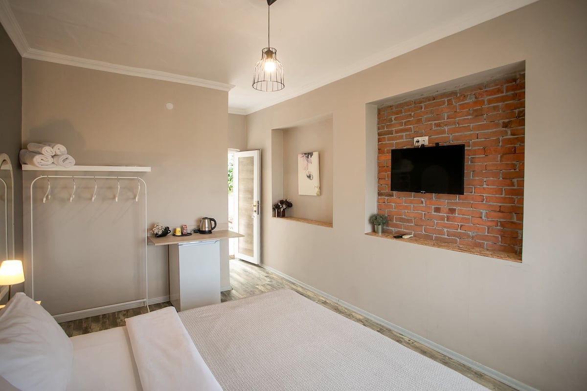 A hotel room with a bed, a desk, a TV mounted on a brick wall, and a hanging light fixture.
