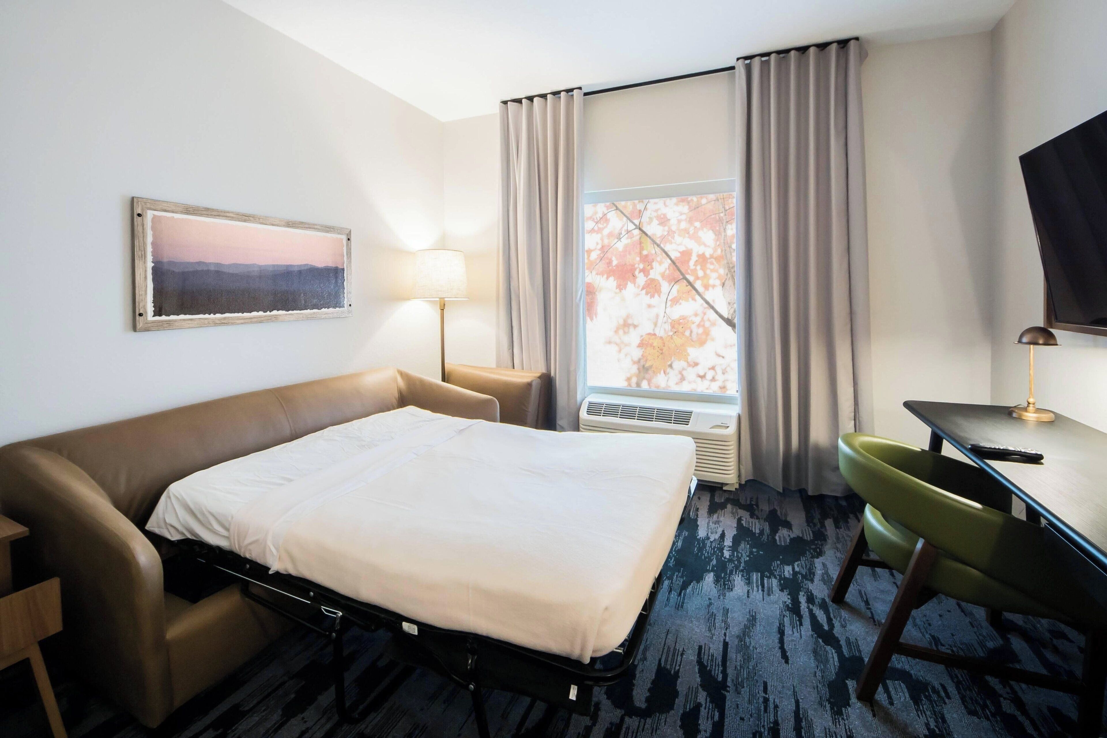 Hotelkamer met een bed, bureau, stoel, televisie, raam met gordijnen en een schilderij aan de muur.