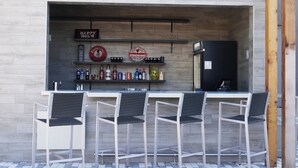 Un área de bar al aire libre con mostrador, taburetes y refrigerador.