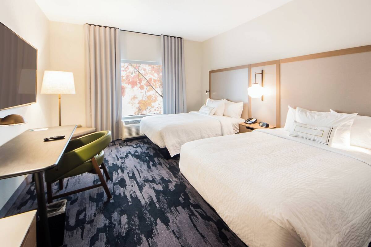 A hotel room with two beds, a desk, a chair, a lamp, and a window with curtains.