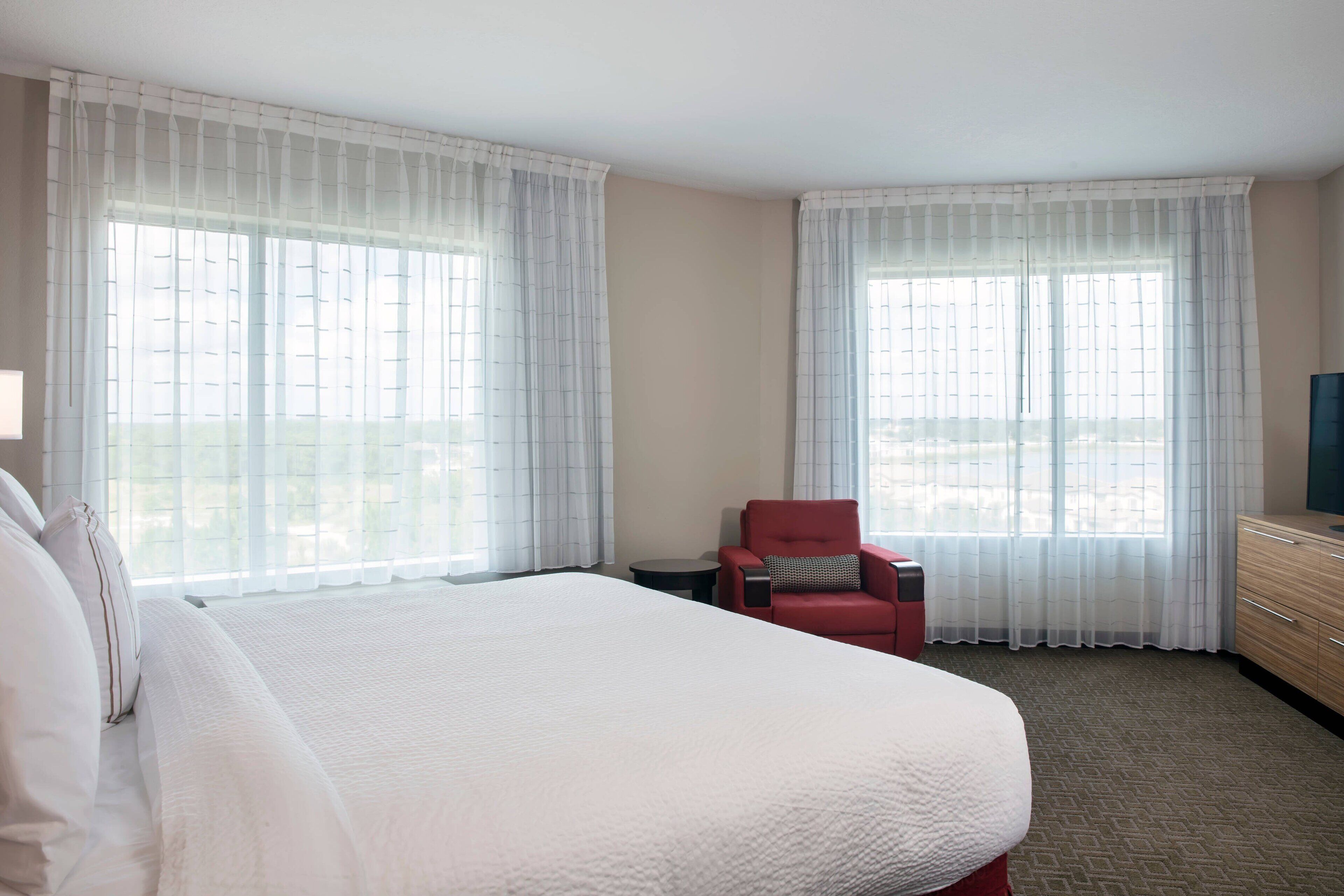 Une chambre d’hôtel avec un grand lit, deux fenêtres avec des rideaux légers, un fauteuil rouge, une petite table et un téléviseur posé sur une commode en bois.