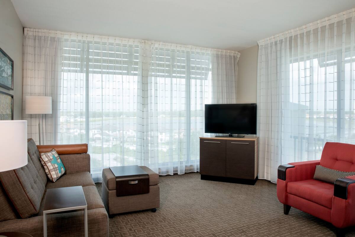 A hotel room with a sofa, ottoman, red armchair, TV, and a cabinet.