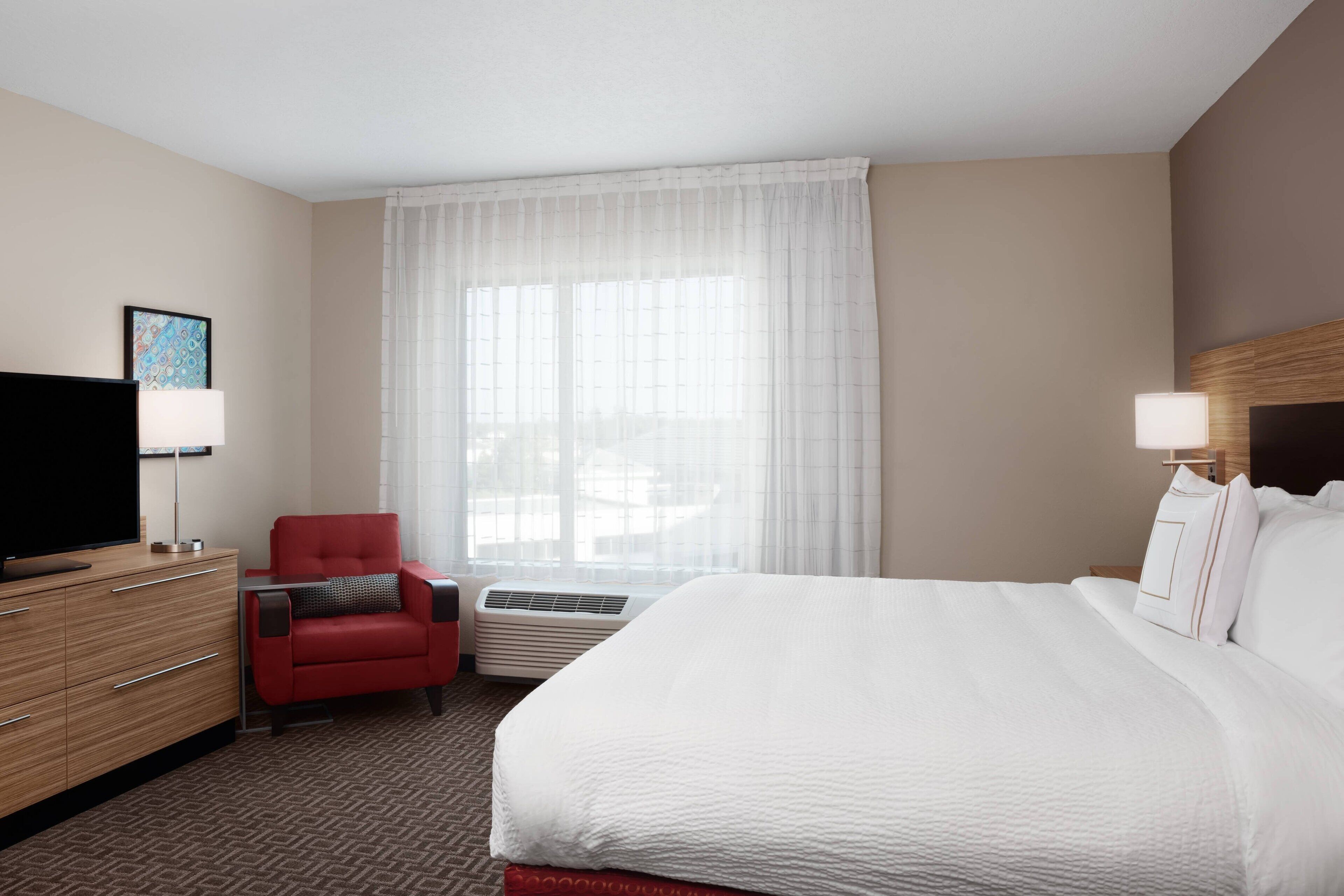 Een hotelkamer met een bed, een rode fauteuil, een televisie, een bureau en een raam met dunne gordijnen.