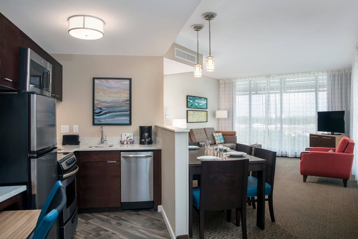 A modern hotel room with a kitchenette, dining area, and a living space with a TV and red armchair.