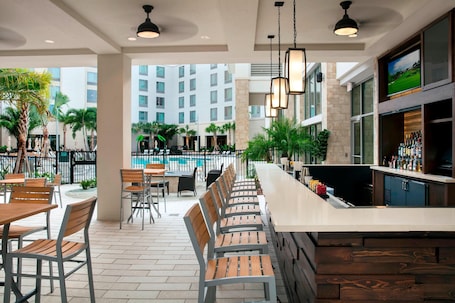 A bar area with wooden chairs and a long counter, overlooking a pool and a building with multiple floors.