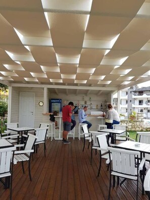 Ein Außenbereich mit einer Terrasse, weißen Tischen und Stühlen sowie einer Bar und einem Holzdeck.