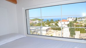 A bedroom with a large window offering a view of a coastal town and the sea.