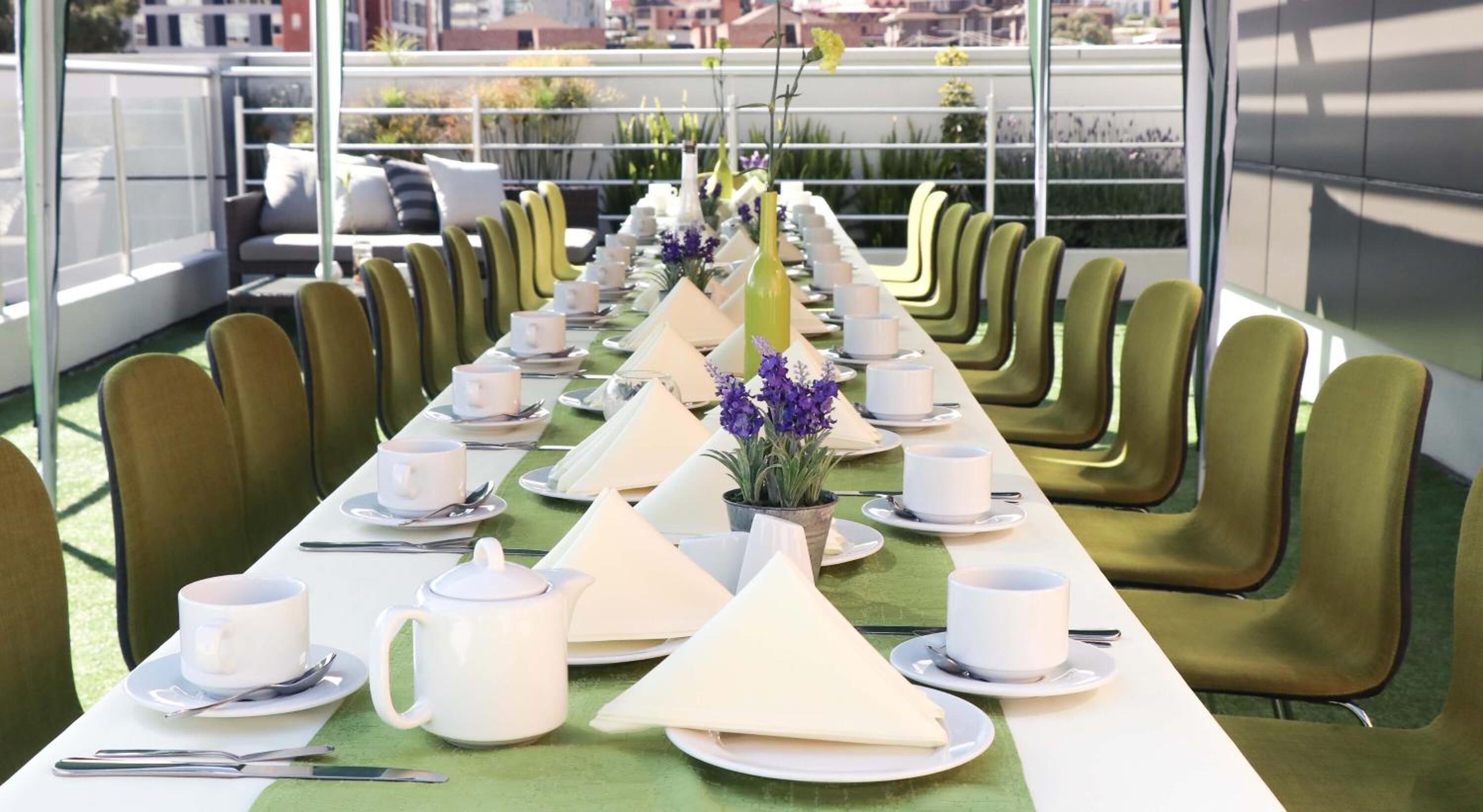 Una larga mesa de comedor con platos blancos, tazas y cubiertos, rodeada de sillas verdes, con vistas a la ciudad al fondo.