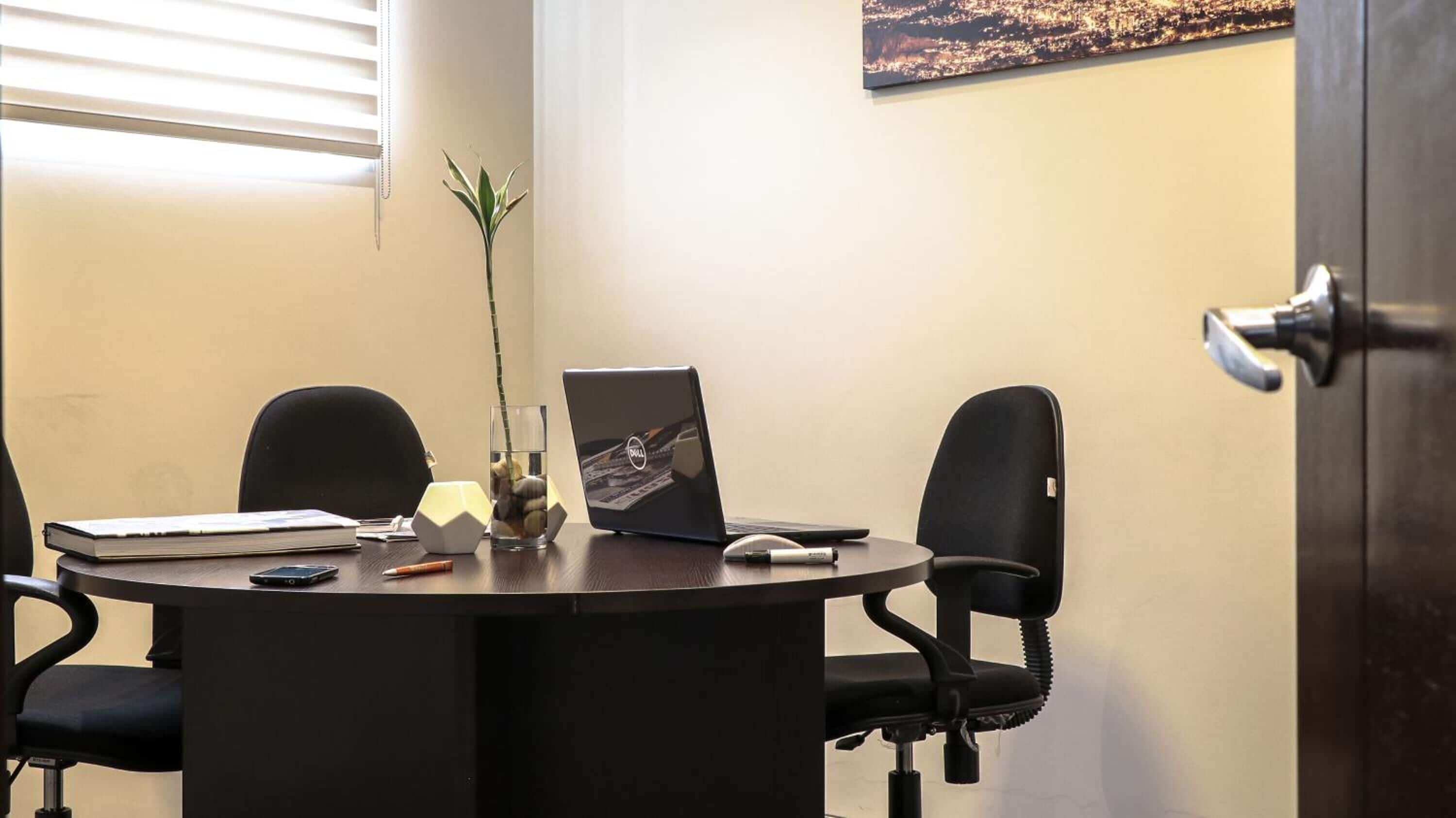 Uma sala de conferências com uma mesa redonda, duas cadeiras pretas, um laptop, um caderno, uma caneta, um copo com gelo e uma planta em vaso.