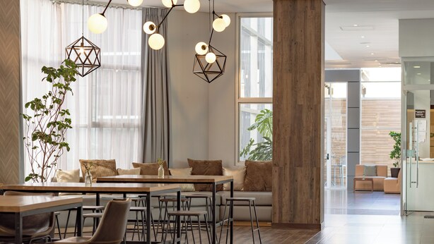 A modern dining area with a wooden table, metal chairs, and a sofa. There are hanging pendant lights and a large window with sheer curtains. - Hotel Rennova (La Paz)