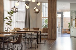 Un espace repas moderne avec une table en bois, des chaises en métal et un canapé. On y trouve des lampes suspendues et une grande fenêtre ornée de rideaux légers.