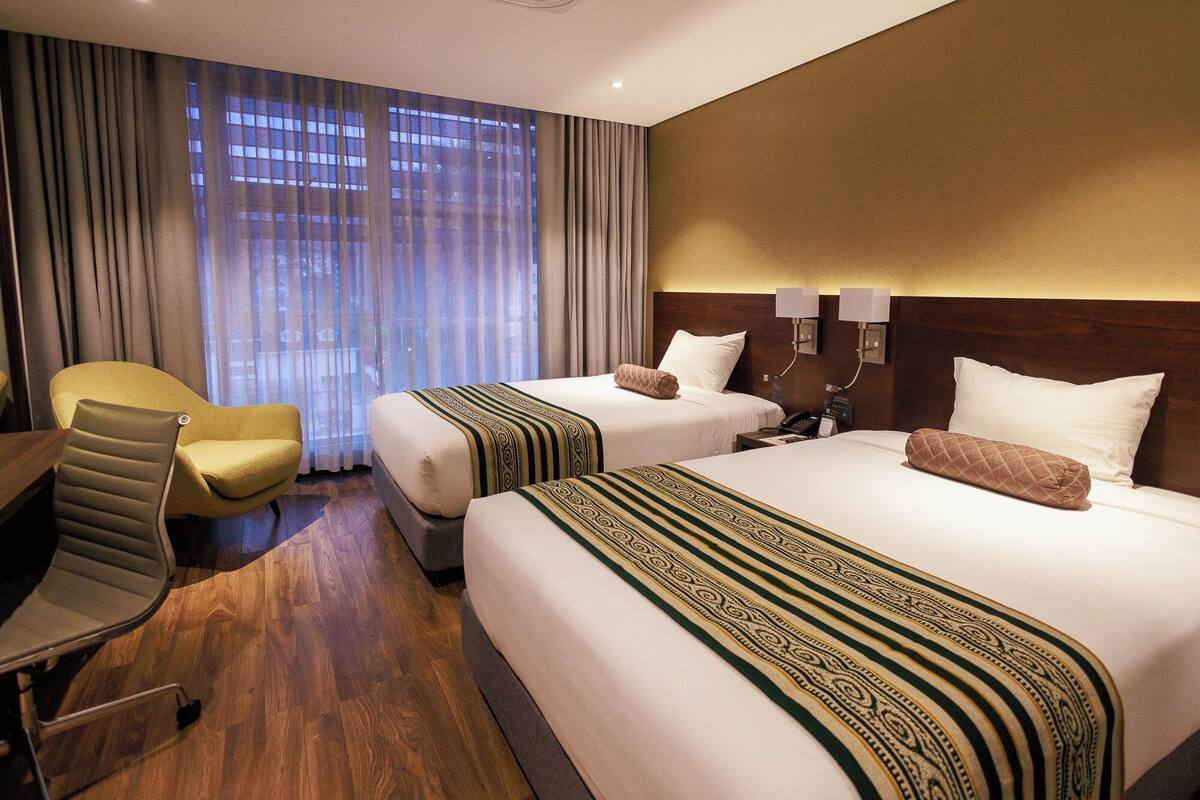 A hotel room with two beds, a desk, a chair, and a large window with curtains.