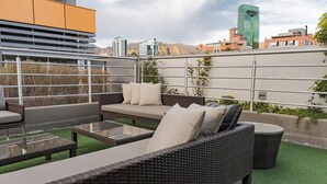 A rooftop patio with wicker furniture, a glass table, and a view of city buildings.