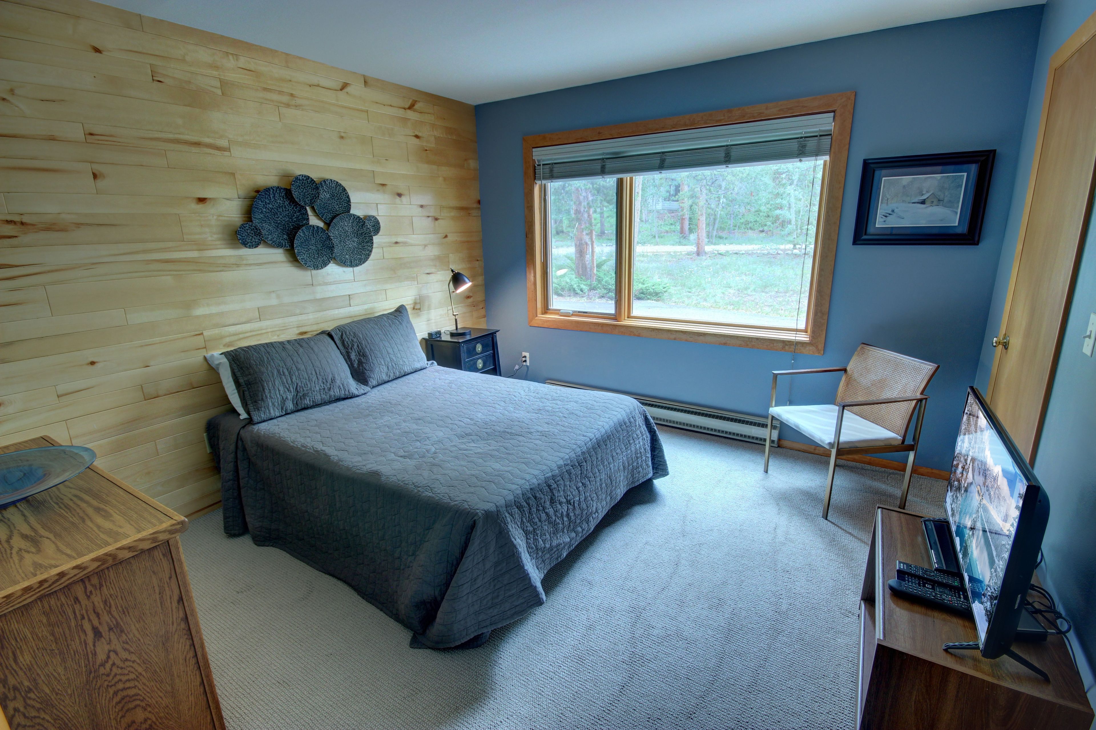A bedroom with a bed, a desk with a computer, a chair, a window with a view of trees, and a wall-mounted decoration.