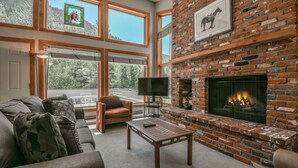 A living room with a brick fireplace, large windows, a sofa, and a television. - Keystone Bristlecone Home 31 (Keystone)