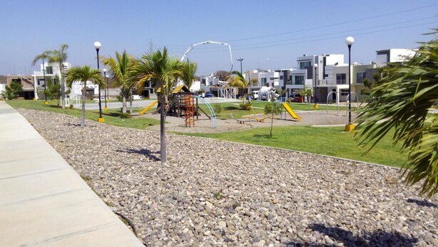 A playground with climbing structures and slides, surrounded by pebbles and palm trees, with modern buildings in the background.