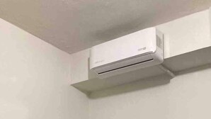 A small, single-bed room with a dark headboard and a grey bedspread. An air conditioning unit is mounted on the wall above the bed.