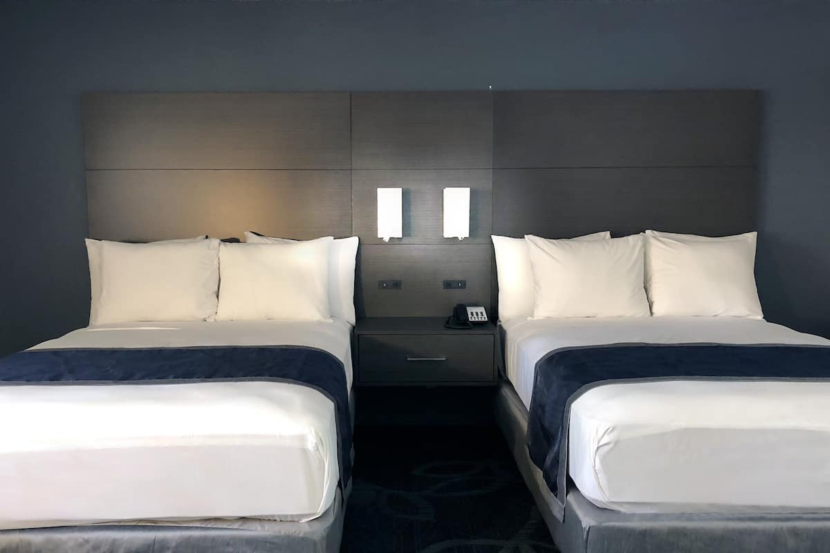A hotel room with two single beds, a nightstand with a phone, and a headboard with built-in lighting.