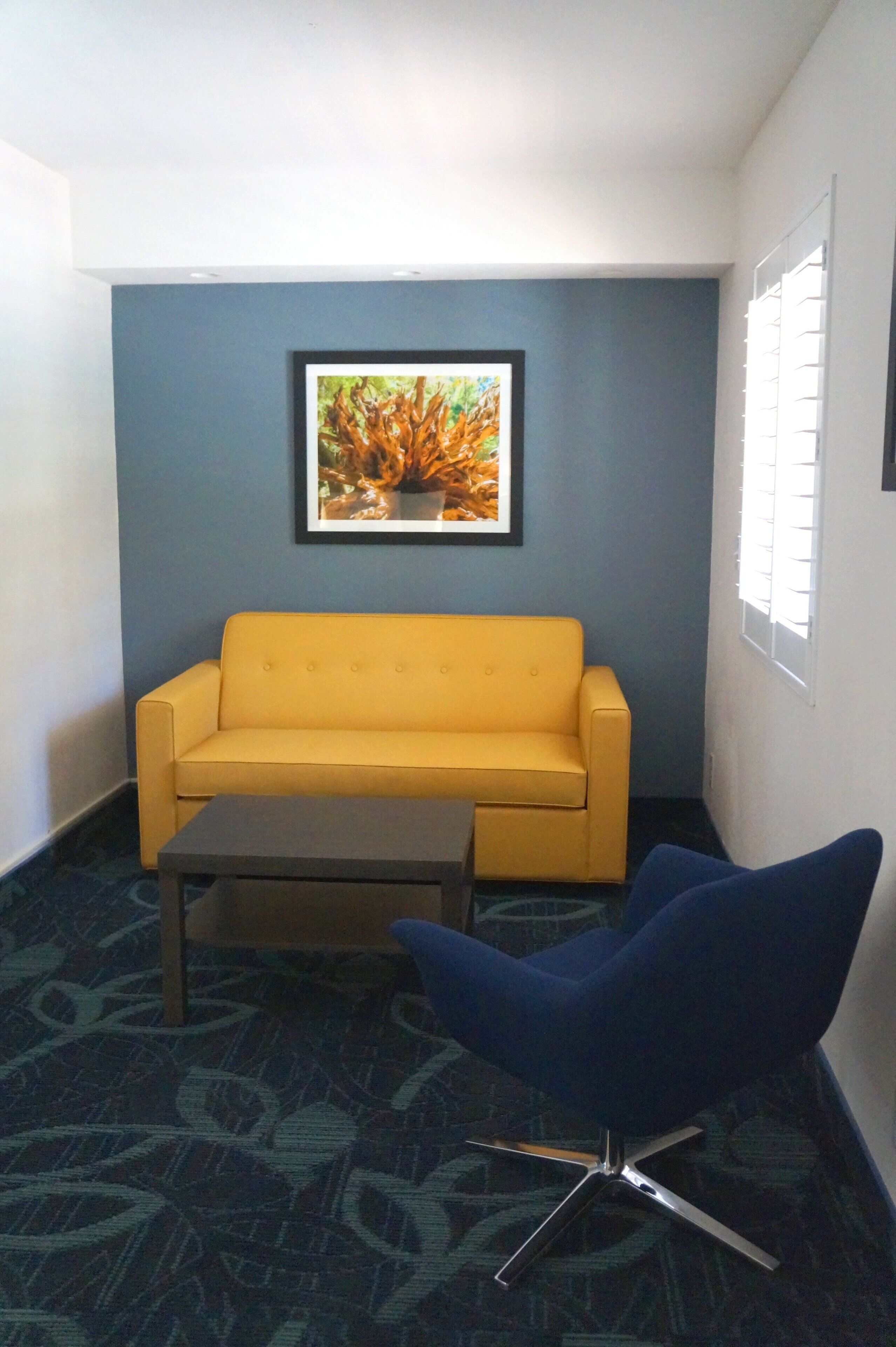 A room with a yellow sofa, a dark coffee table, and a blue chair. A framed picture hangs on the wall.