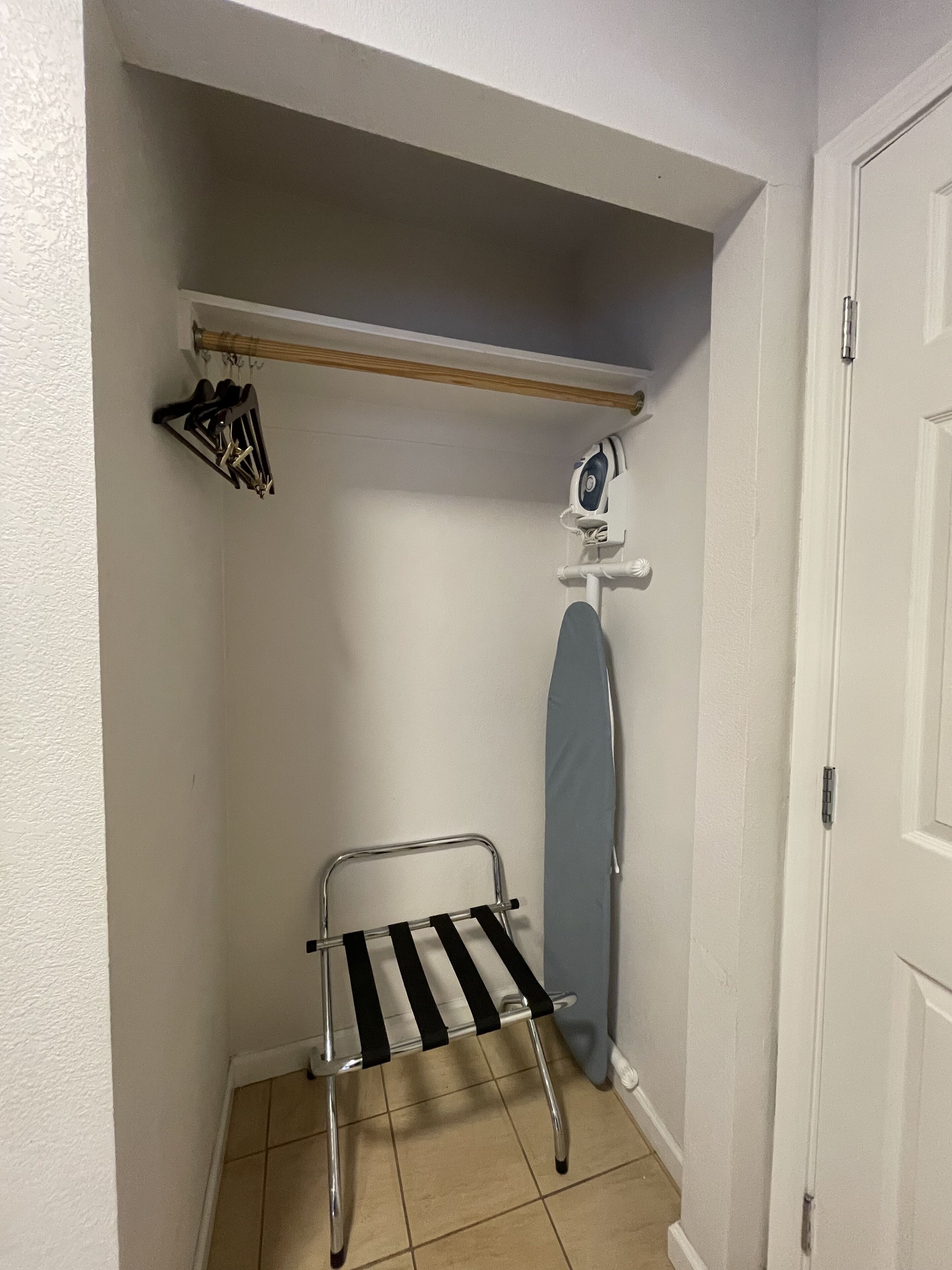 A small, narrow room with a metal drying rack, a wall-mounted ironing board, and a shelf with hanging items.