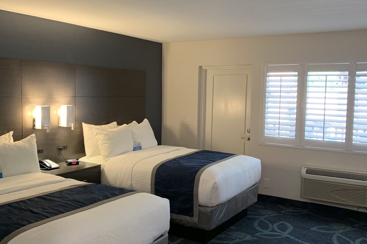 A hotel room with two beds, bedside lamps, a nightstand, a phone, and a window with blinds.
