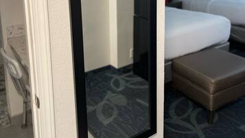 Quarto de hotel com um espelho grande, uma cama com roupa de cama branca, um banquinho marrom e uma cabeceira com painel escuro.