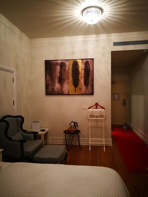 A hotel room with a bed, a chair, a small table with a kettle, a coat rack, and a painting on the wall. - Heritage Apartment (Auckland)