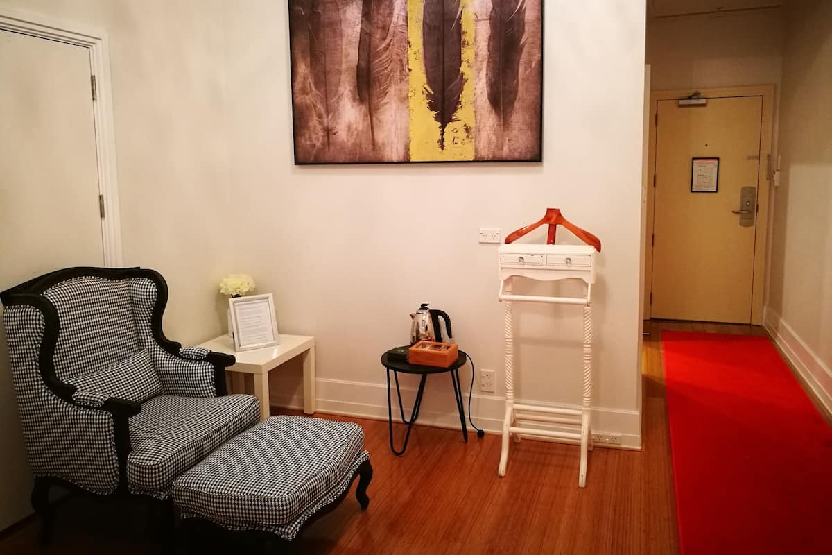 A room with a checkered armchair, a small table with a vase and a framed document, a white side table, a coat rack, and a red carpet leading to a door.
