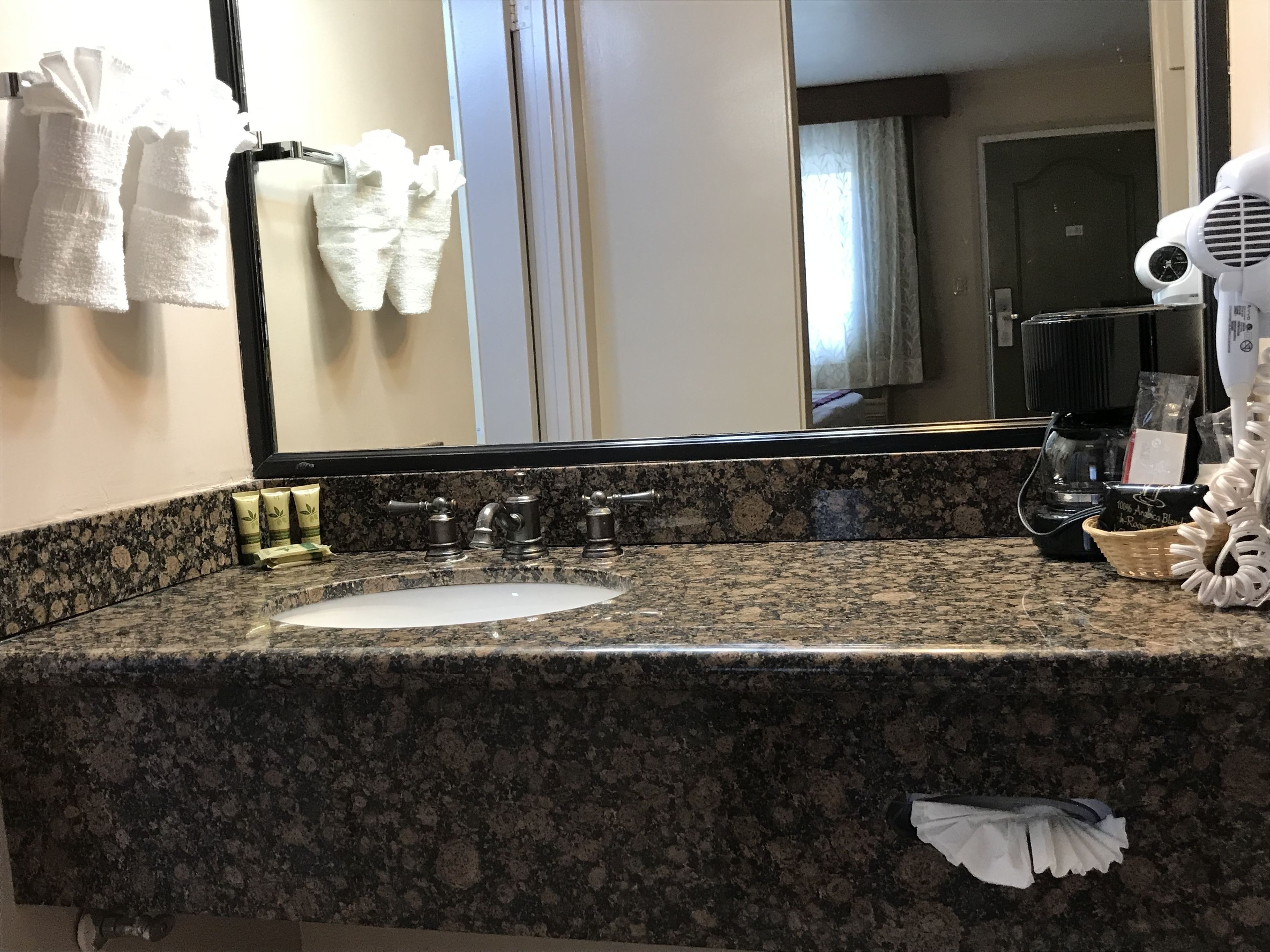 A hotel bathroom with a granite countertop, a sink, a mirror, a hairdryer, and a coffee maker.