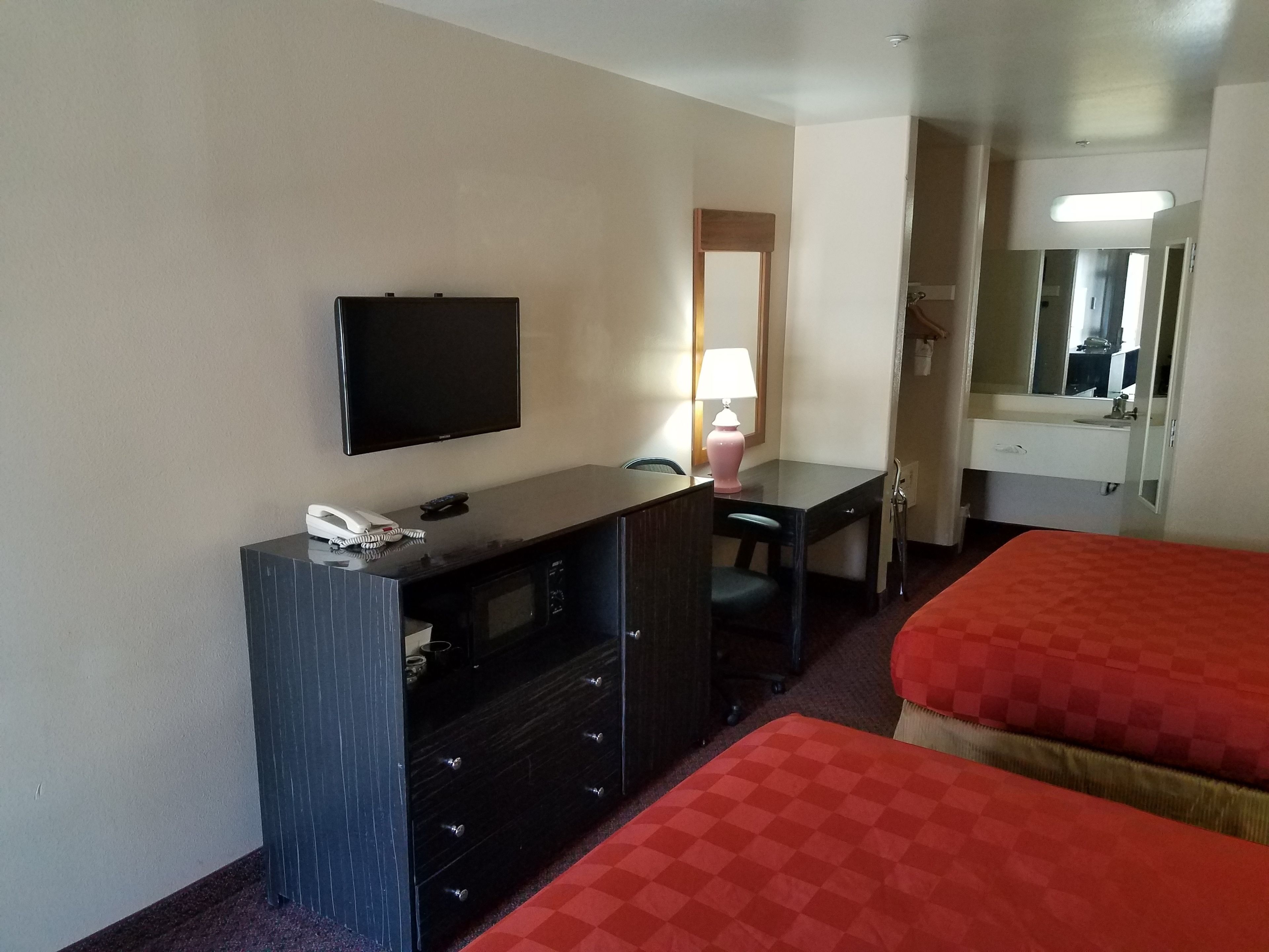 Ein Hotelzimmer mit Flachbildfernseher, Schreibtisch mit Lampe, Telefon und Minibar. Es gibt zwei Betten mit roter Bettwäsche.