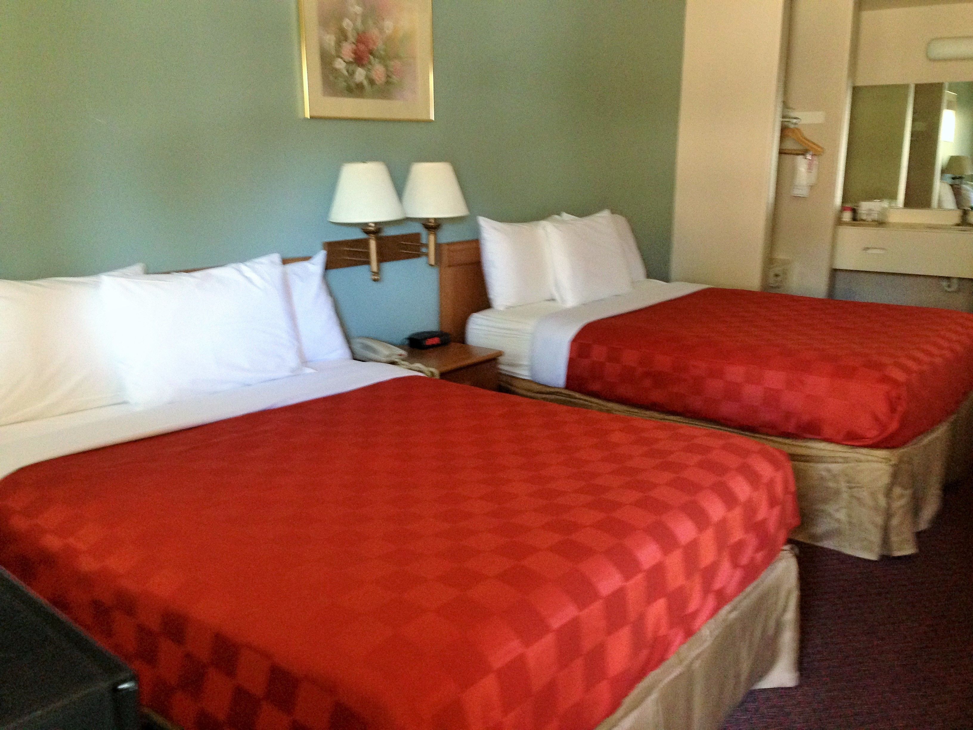 Habitación de hotel con dos camas, cada una con ropa de cama blanca y colchas rojas. Hay una mesita de noche con una lámpara y un teléfono entre las camas. Un cuadro cuelga en la pared sobre las camas.