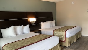 Une chambre d’hôtel avec deux lits, chacun doté de draps blancs et d’une couvre-lit beige et rouge. Il y a une table de chevet entre les lits, une lampe fixée au mur au-dessus de la tête de lit et un plancher en bois.