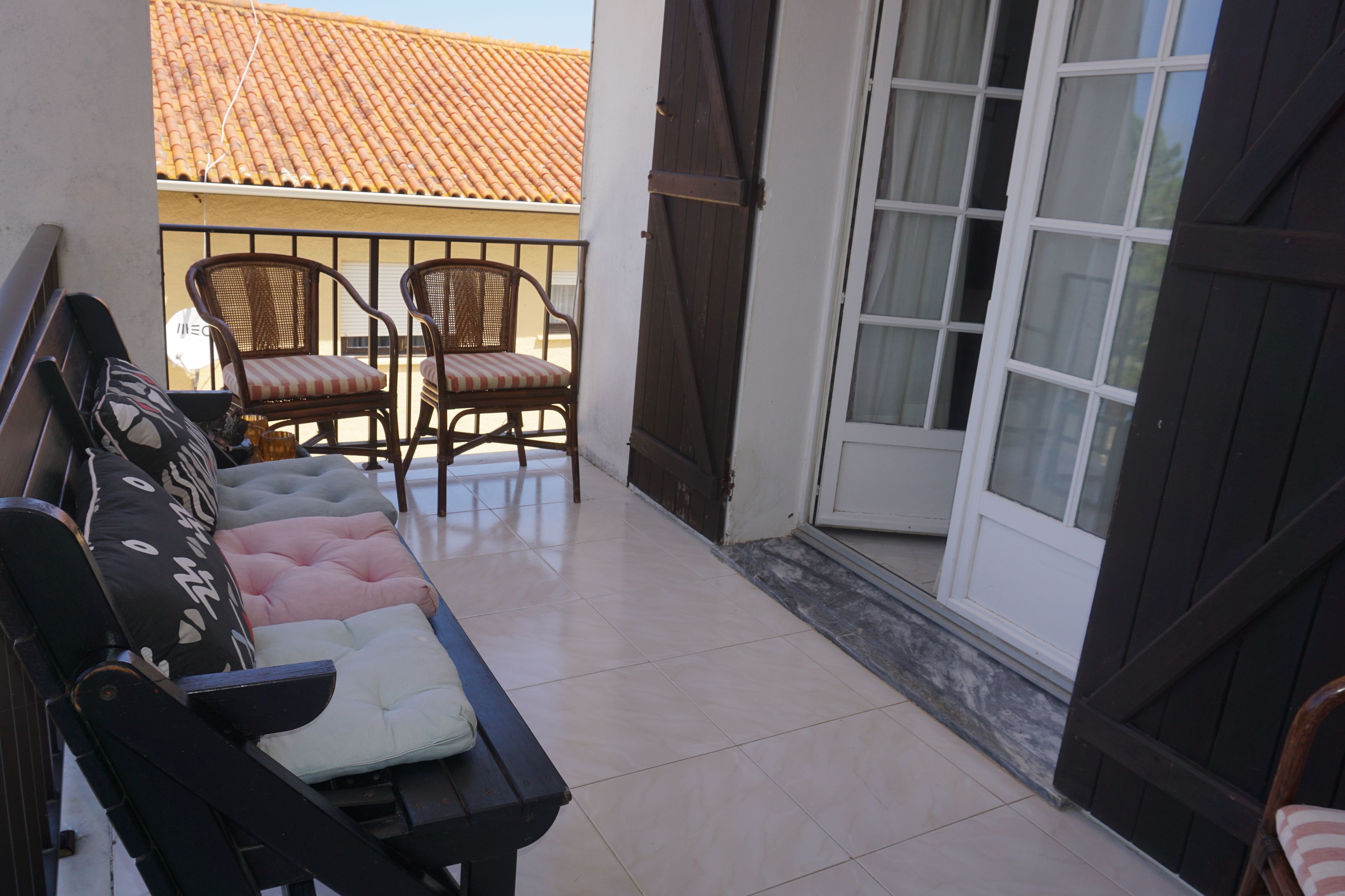 A balcony with wicker chairs, a cushioned bench, and a sliding glass door leading to a tiled room.