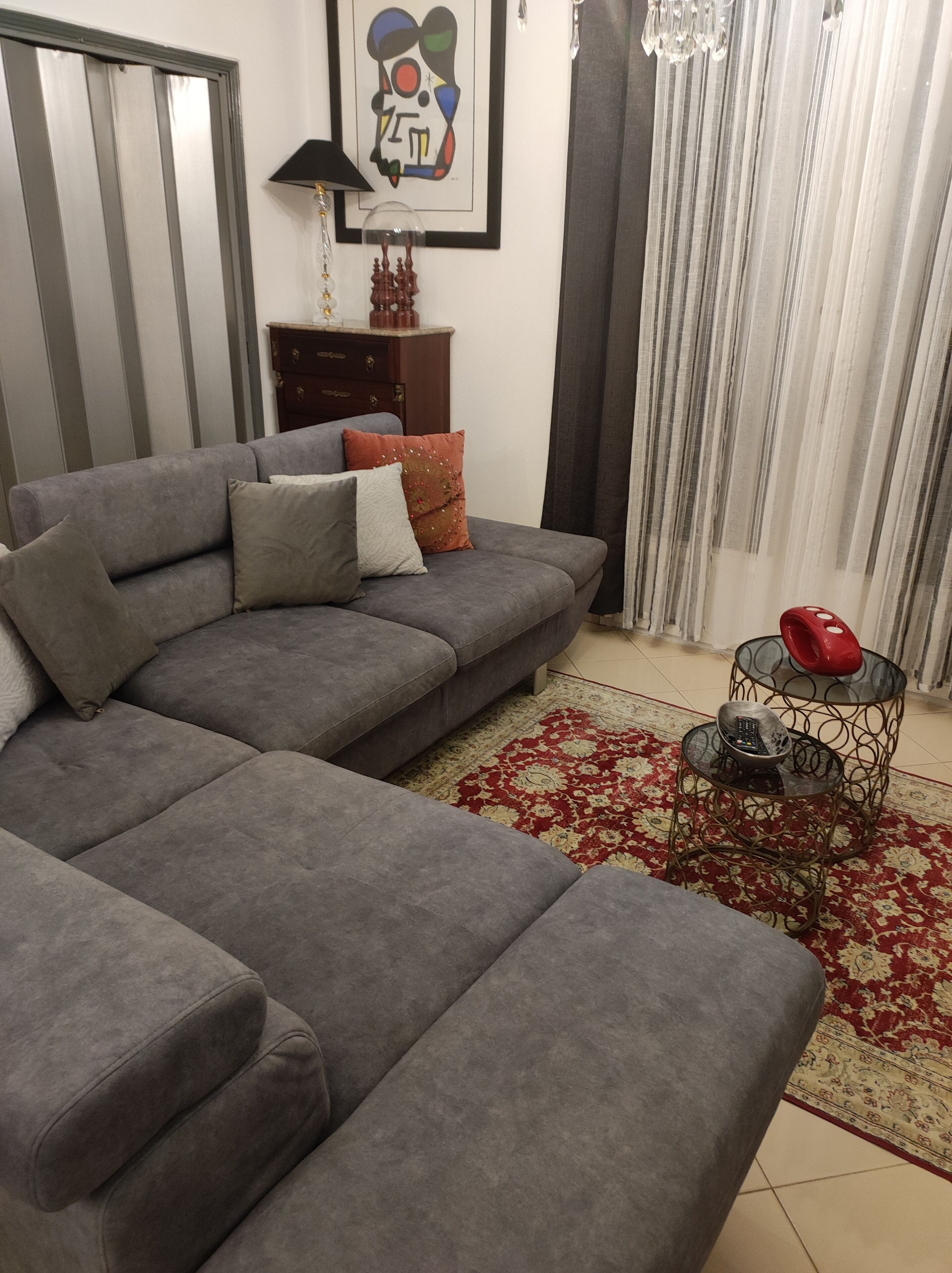 A modern living room with a grey sectional sofa, a red patterned rug, a glass coffee table with a red object, and a framed abstract painting on the wall.