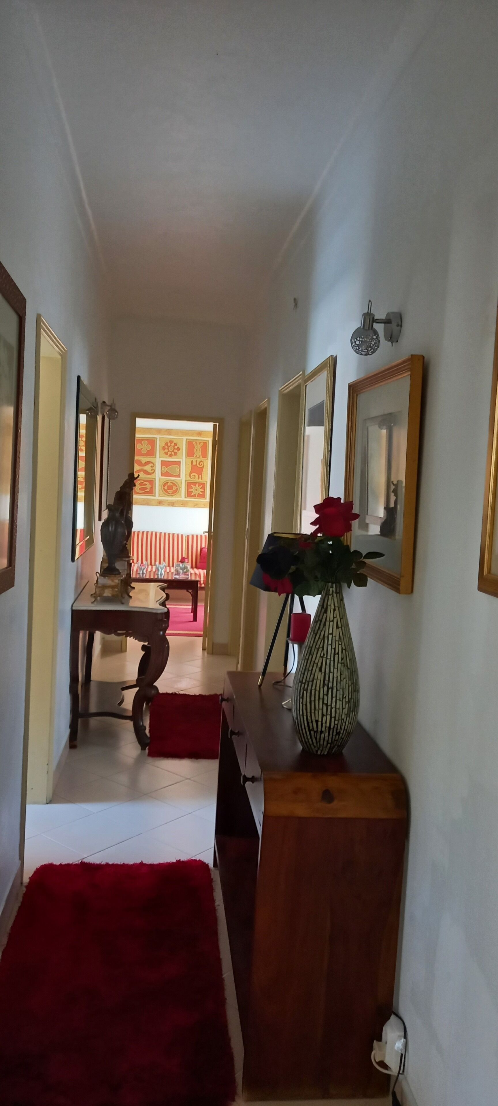 Um corredor com carpete vermelho, um console de madeira com um vaso de flores e um espelho na parede.