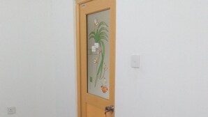 Uma porta com um painel de vidro e fechadura, um tapete vermelho no chão, e uma parede branca com um interruptor de luz.