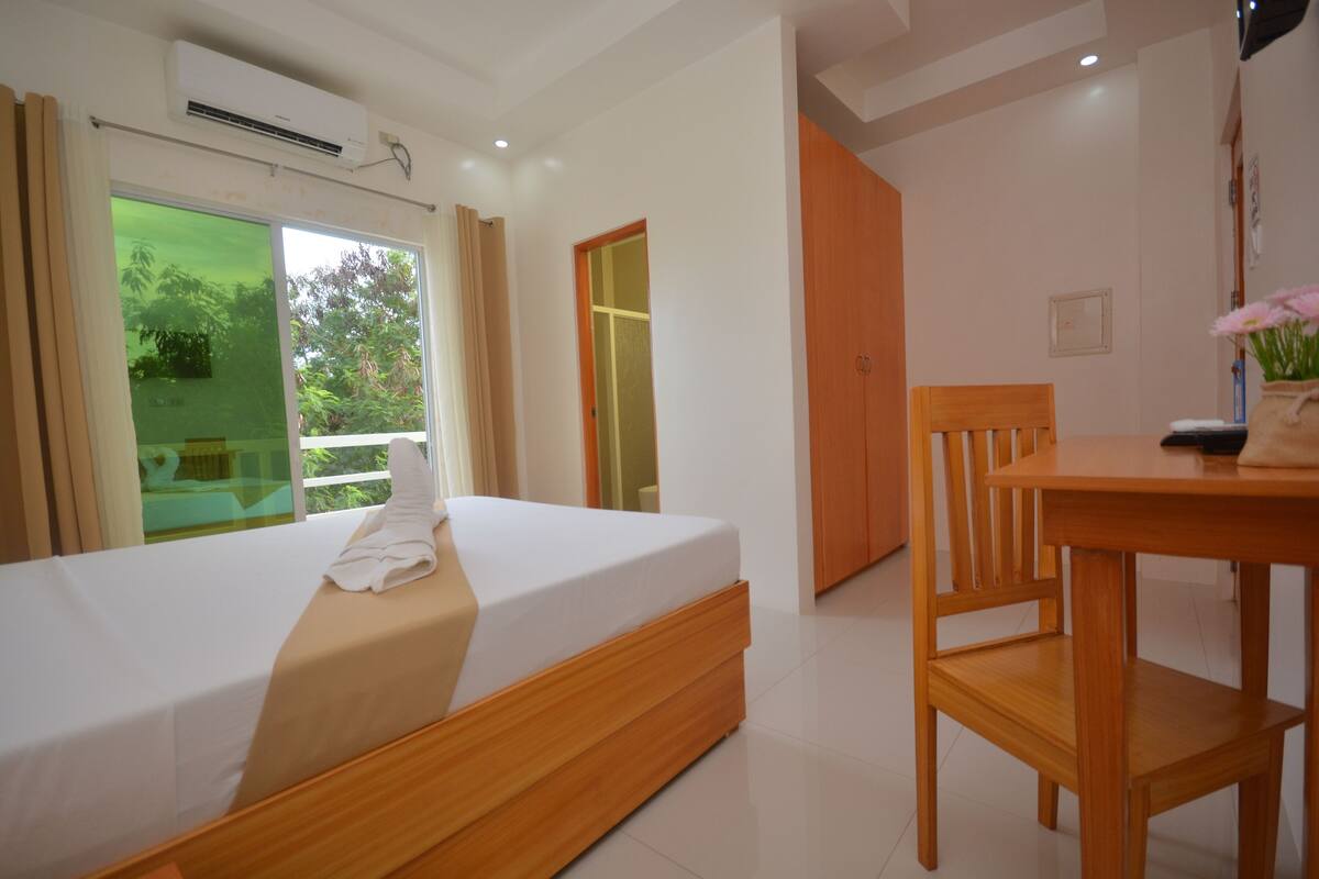 A hotel room with a single bed, a wooden chair, a table, and a view of greenery through the window.