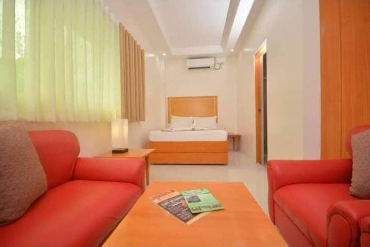 A hotel room with a bed, two red sofas, a wooden coffee table, and a window with sheer curtains.
