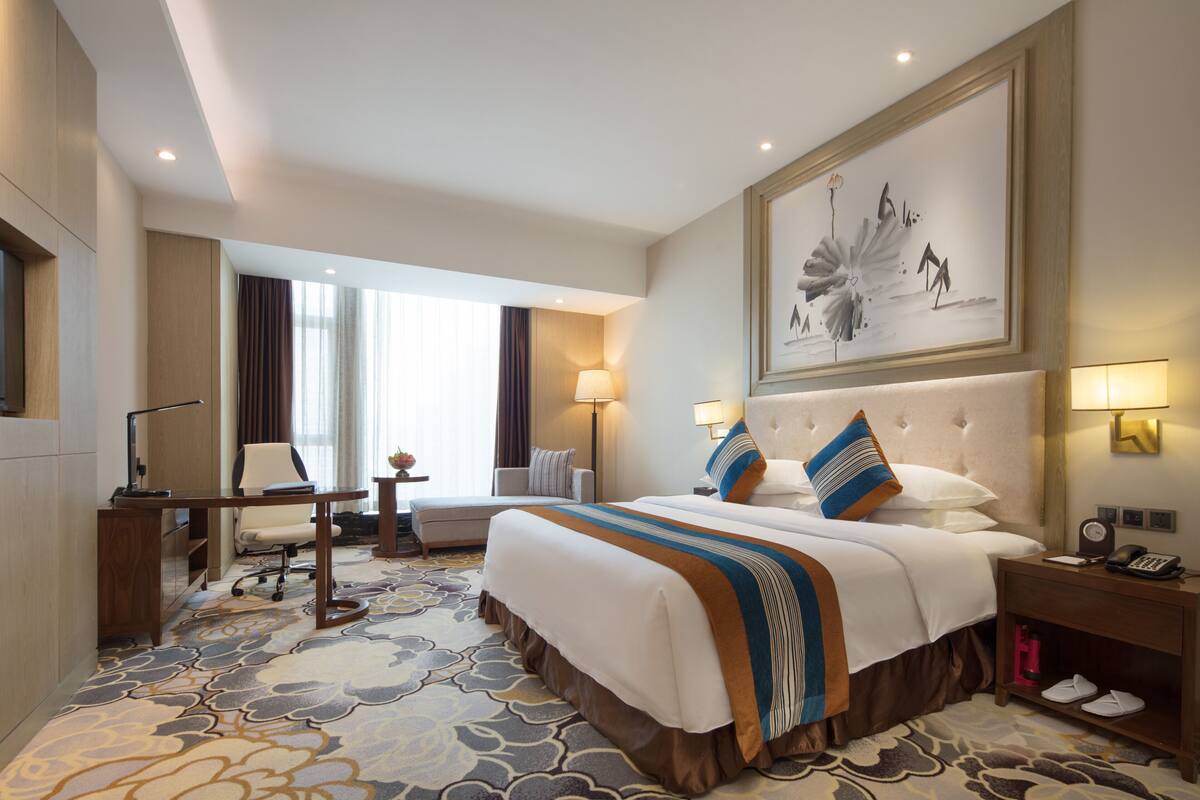 Quarto de hotel com uma cama grande, uma escrivaninha, uma cadeira e uma janela ampla.