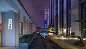 Vista nocturna de un pasillo de hotel con un edificio moderno a la derecha, un camino bien iluminado y un macetero decorativo.