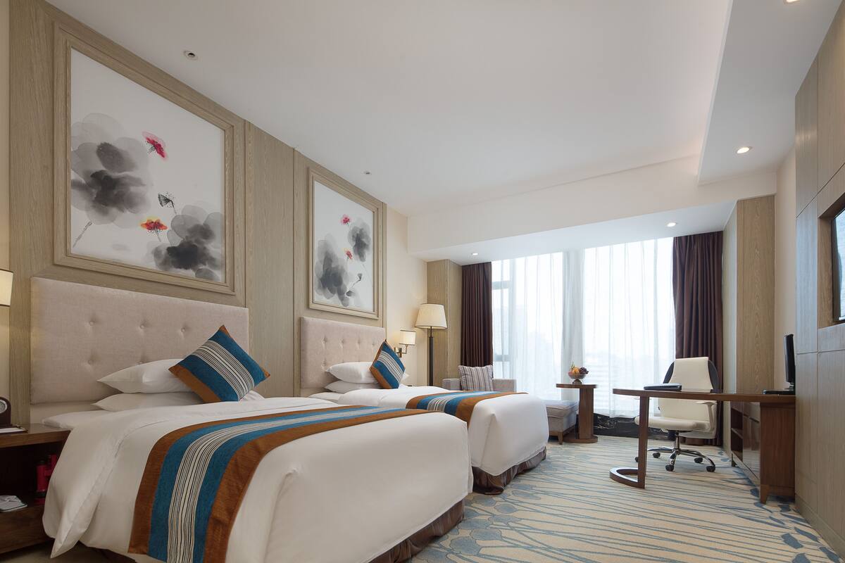 Quarto de hotel com duas camas, uma escrivaninha, uma cadeira, televisão e obras de arte nas paredes.